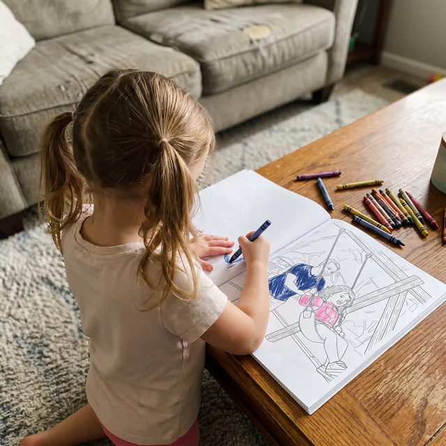 A little girl kneeling at a coffee table, coloring the page of her and her dad on the swing